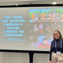 Alex Piscatelli stands next to a screen with the title of her presentation: The Tribe Has Spoken: Fandom & Social Change on Survivor